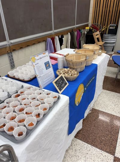 A table display features trays of small white cups with food items, informational signs, and wicker baskets.